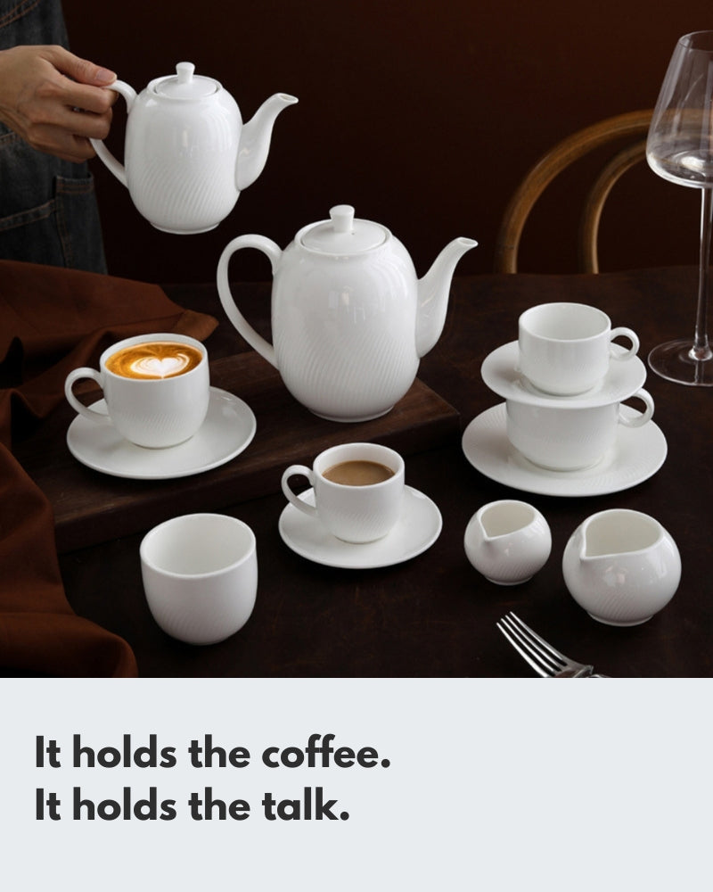 An elegant white ceramic coffee and tea set with a subtle ribbed texture. The set includes two teapots, two coffee cups (one with latte art), and various saucers and creamers, arranged on a dark wooden table to showcase a sophisticated coffee experience.